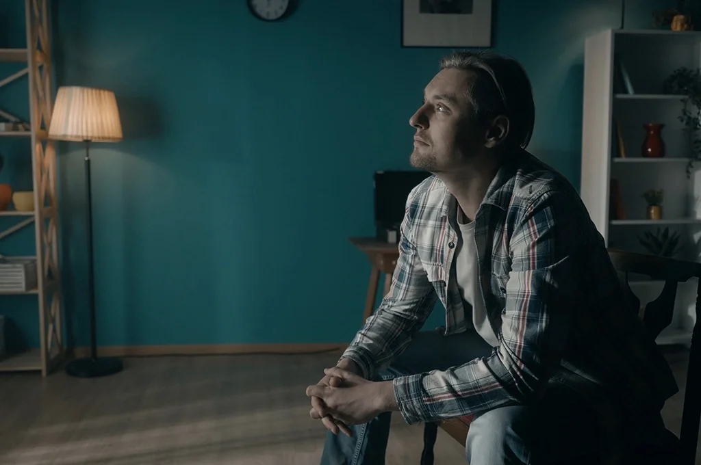 A man sits on a couch and stares out a window with a lamp glowing in the background